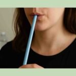 Close-up of a young woman’s lower face and hand practicing straw phonation with a long blue straw, wearing a black shirt, with blurred sheet music and a water bottle in the background.