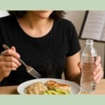 Singer holding a water bottle in one hand and a fork poised over a plate of grilled chicken, brown rice, and avocado, with a blurred sheet music stand in the background.