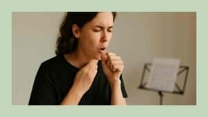 Singer in a black T-shirt clears her throat, hand on her throat, against a neutral background.
