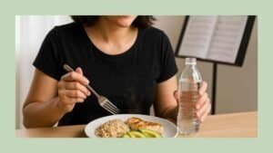 Singer holding a water bottle in one hand and a fork poised over a plate of grilled chicken, brown rice, and avocado, with a blurred sheet music stand in the background.