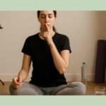 Singer seated cross-legged on a lavender yoga mat, eyes closed in meditation, finger to lips for vocal rest, with a music stand, bottled water, and rolled yoga mat in the background.