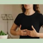 Singer standing with hands on her ribcage, practicing diaphragmatic breathing, with a glass of water, spinach, banana, and nuts on a wooden table and a blurred music stand in the background.