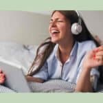 Singer in a rust-colored sweater with curly brown hair, eyes closed, singing into a black microphone while her hand rests on her chest and a music stand is blurred behind her.
