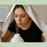 Young woman steaming her face over a humidifier while holding a water bottle, with a towel draped over her head and blurred sheet music in the background.